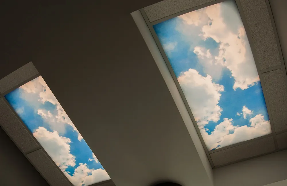Looking up at two rectangular ceiling light panels designed to look like a bright blue sky with fluffy white clouds