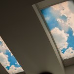 Looking up at two rectangular ceiling light panels designed to look like a bright blue sky with fluffy white clouds
