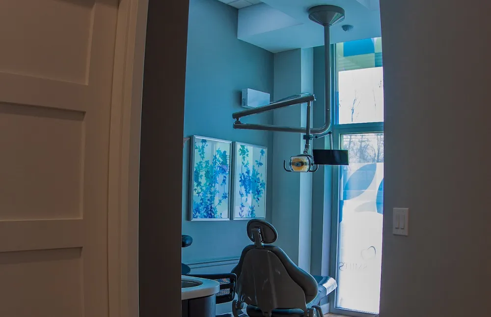 A view into a dental operatory through a doorway, showing a patient chair, blue wall art, and professional overhead lighting