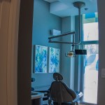 A view into a dental operatory through a doorway, showing a patient chair, blue wall art, and professional overhead lighting