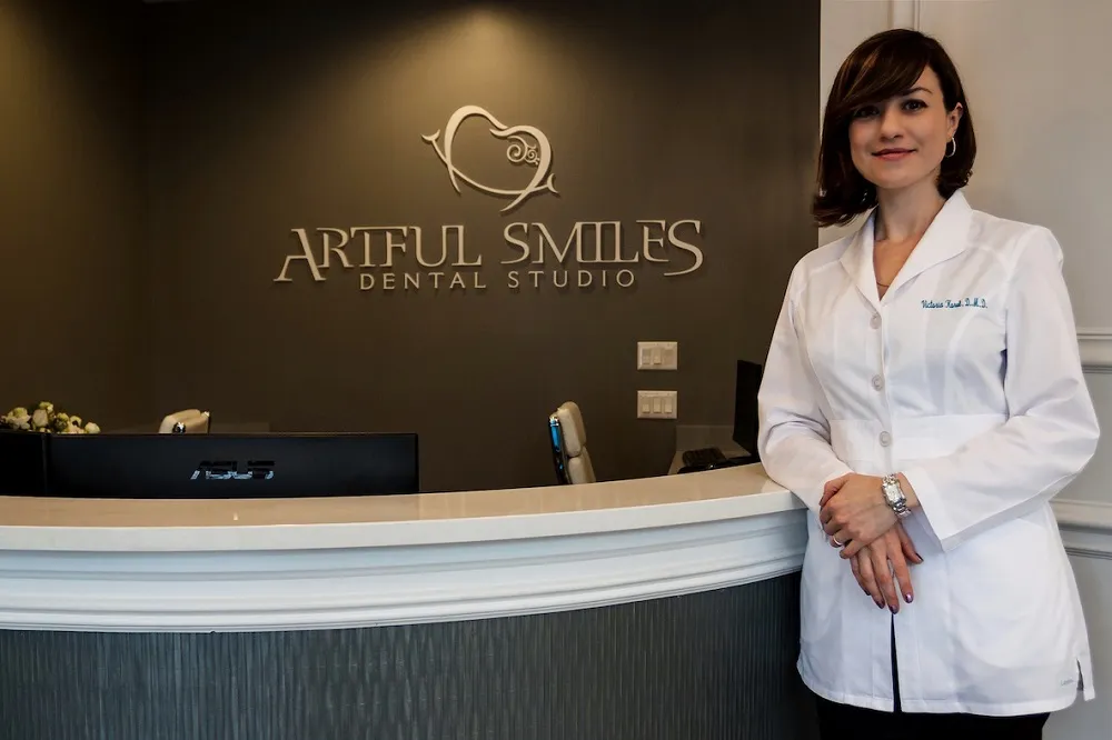 A female dentist in a white lab coat standing confidently next to the curved reception desk in the clinic lobby.