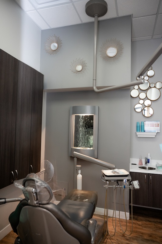 A modern dental room with a gray chair, tall storage cabinets, a large mirror, and decorative wall accents