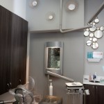 A modern dental room with a gray chair, tall storage cabinets, a large mirror, and decorative wall accents