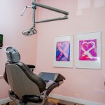 A dental treatment room with soft pink walls, a comfortable patient chair, and colorful abstract art pieces